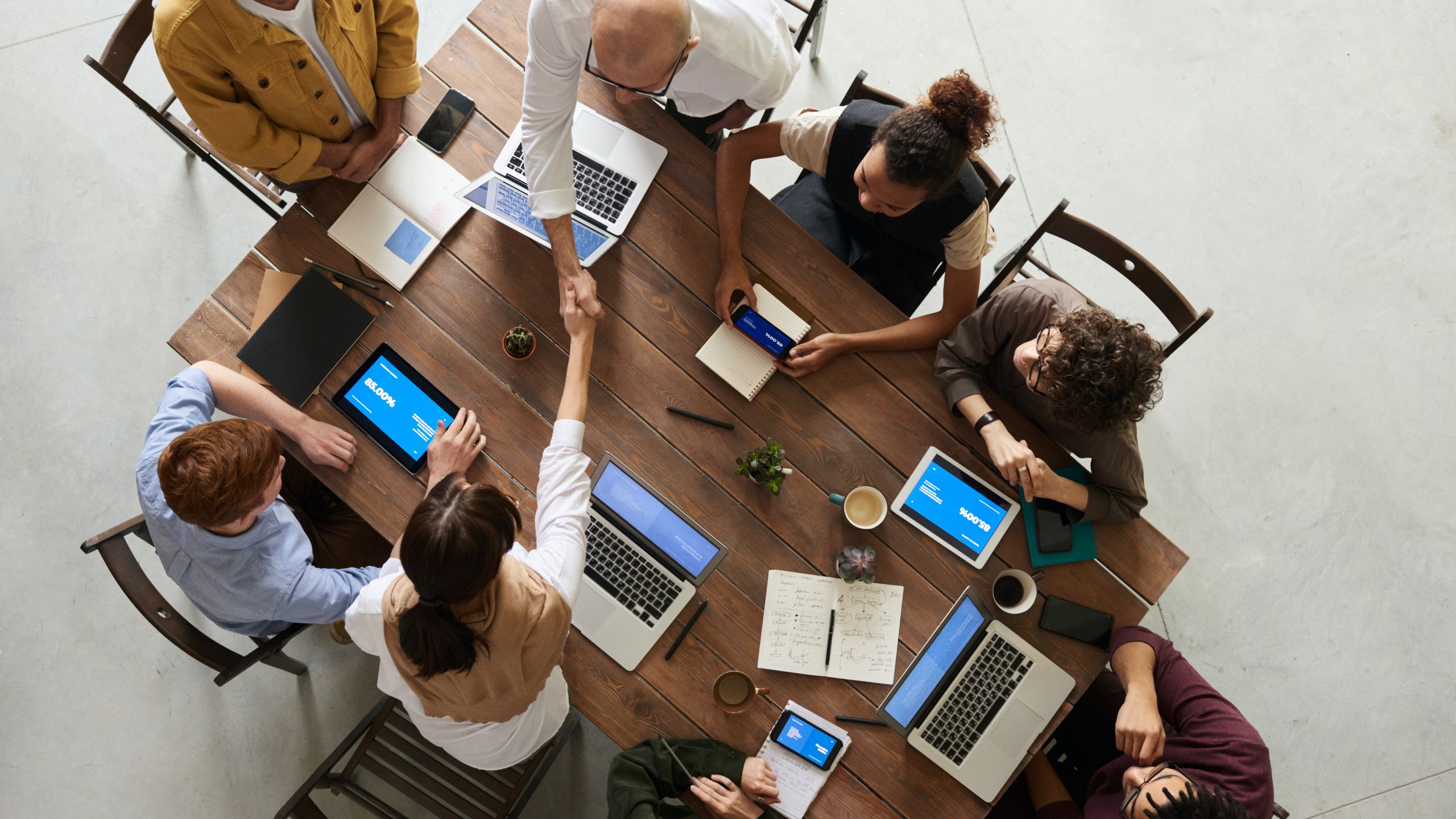 Photo of people sitting around a table with devices. Two people shake hands.